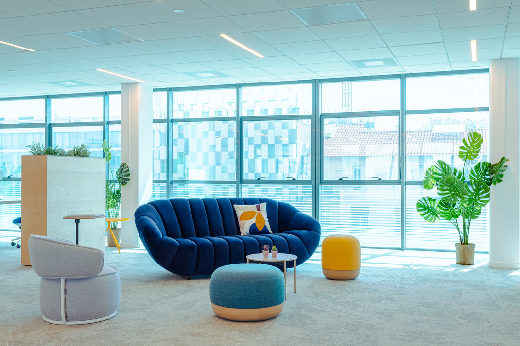 Modern office space with a blue sofa, yellow ottoman, and large windows.