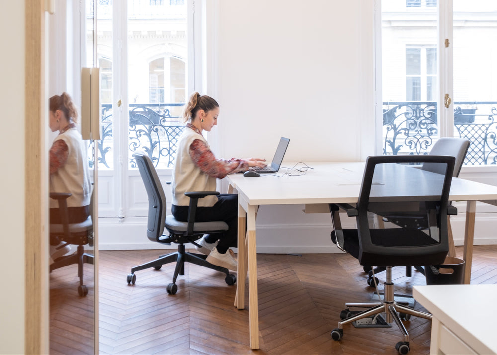 Person working on a laptop at a desk in a modern office setting with large windows.