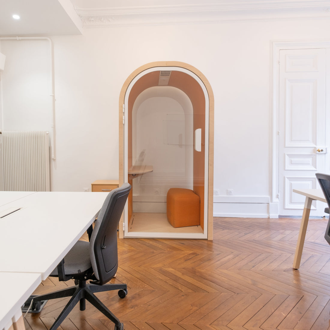 Modern office space with a large desk, chairs, and a small orange office pod.