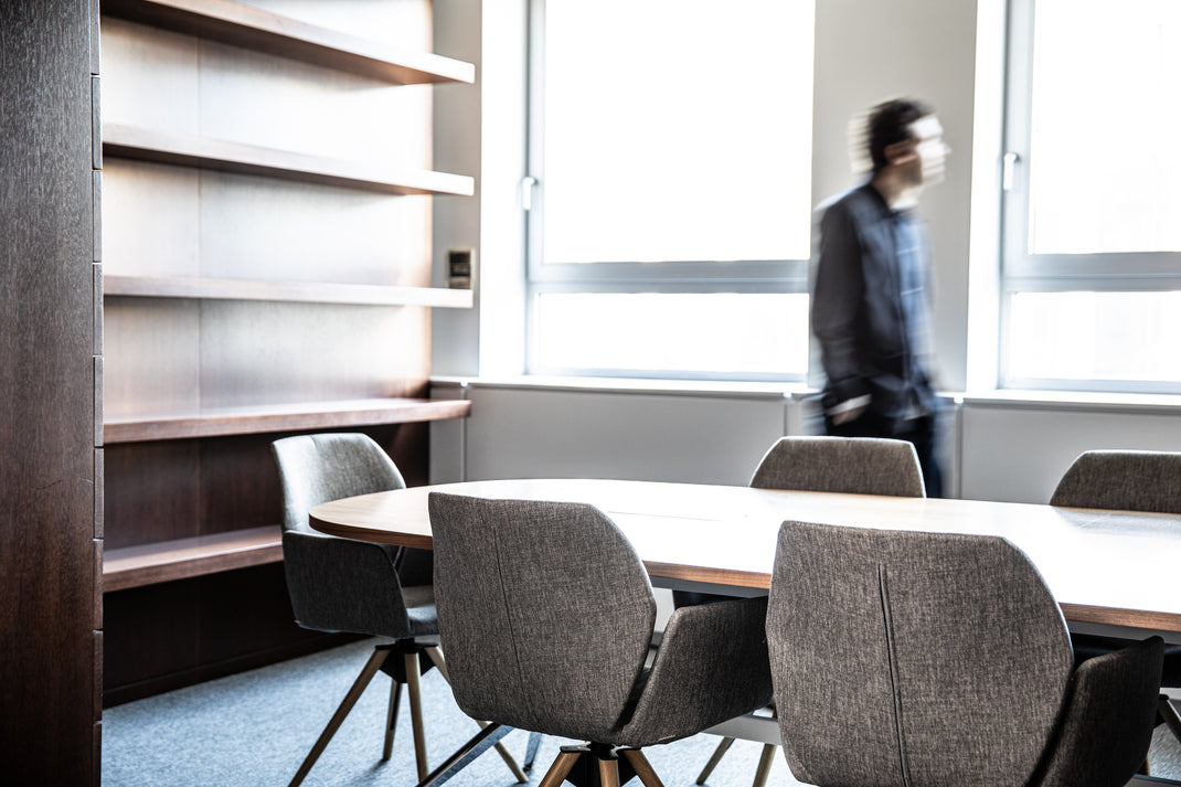 Modern office conference room with round table and chairs, bookshelf, and person in the background.