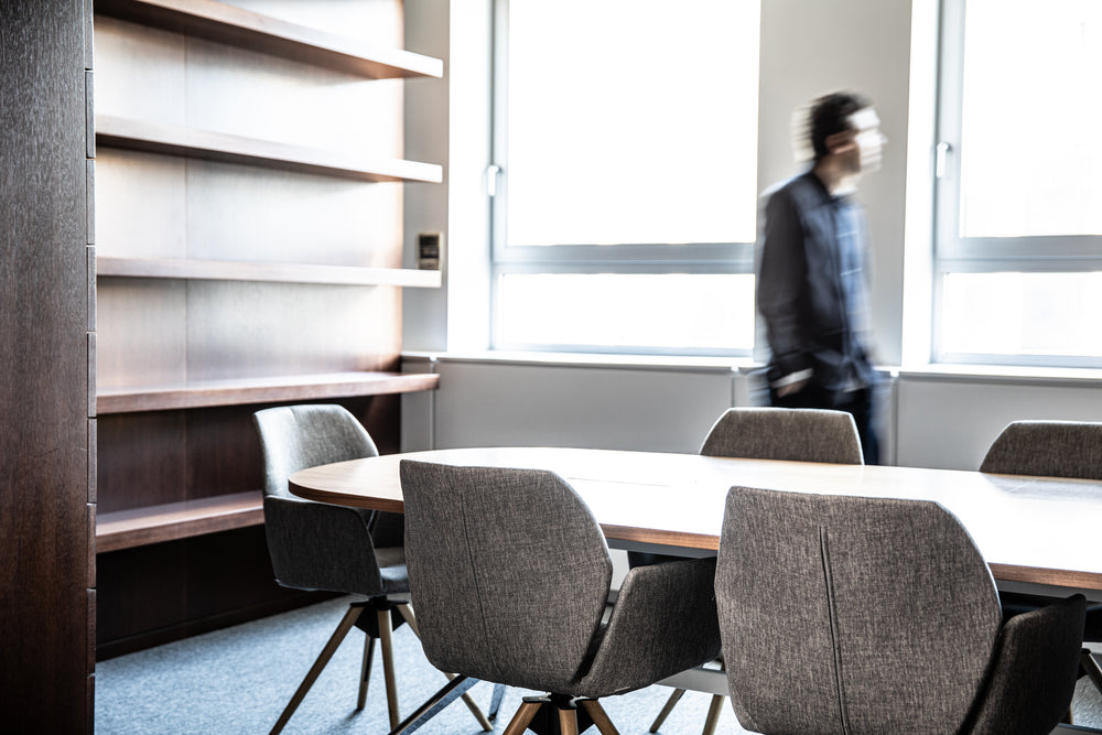 Modern office conference room with round table and chairs, bookshelf, and person in the background.