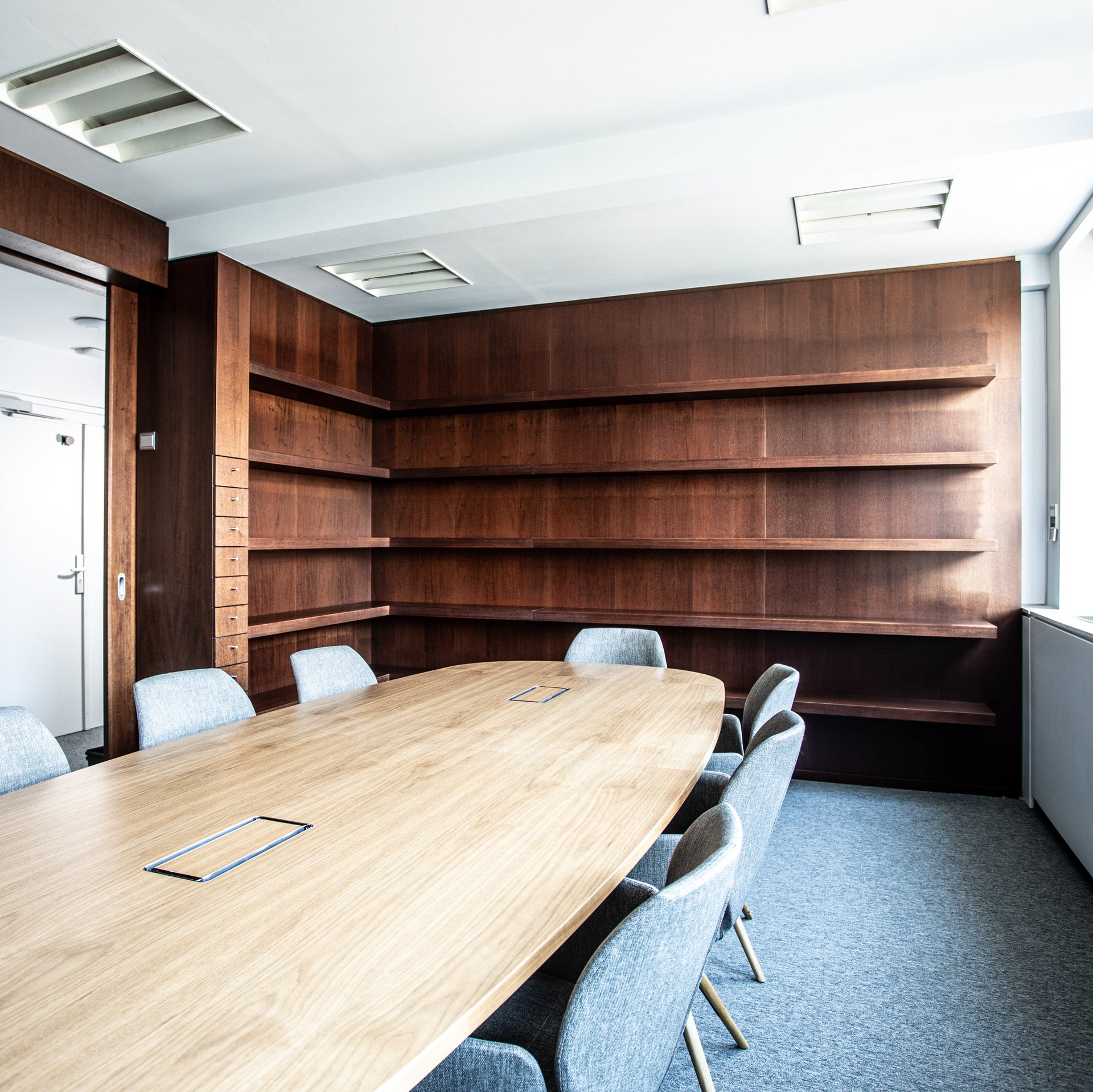 Modern conference room with wooden table and chairs, large bookshelf, and large window.