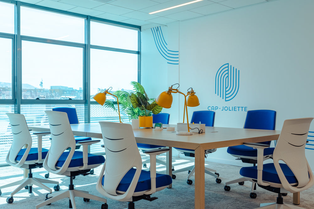 Modern office conference room with blue and white chairs, a large window, and a visible logo on the wall.