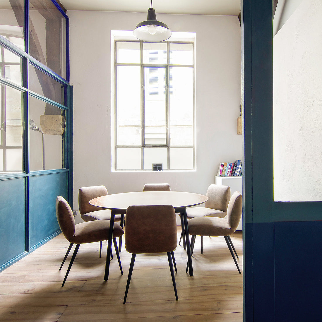 Open office meeting break out zone with round wooden table and leather chairs in a room with large windows.