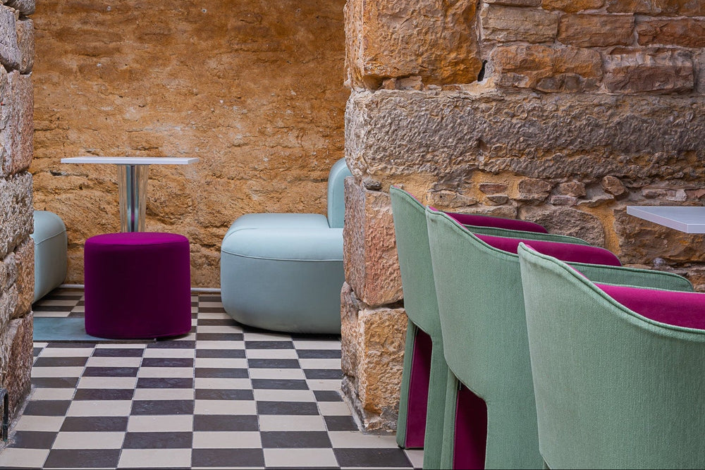 Modern café/restaurant interior with stone walls, checkered floor, and colourful chairs in green, pink and blue.