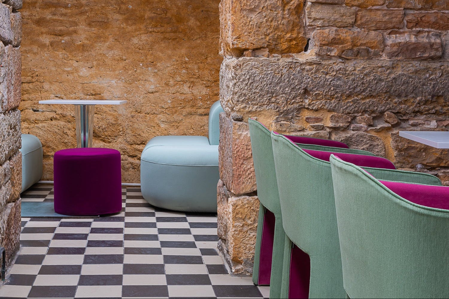 Modern café/restaurant interior with stone walls, checkered floor, and colourful chairs in green, pink and blue.