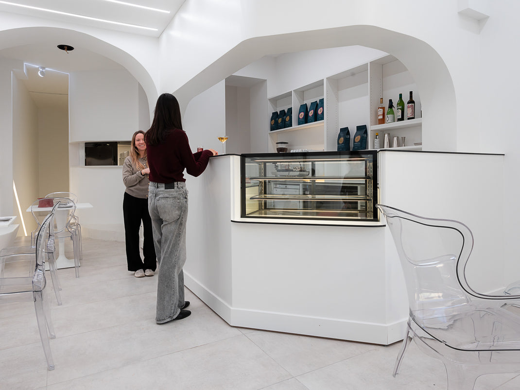 Two women in a modern kitchen with white walls and clear chairs.