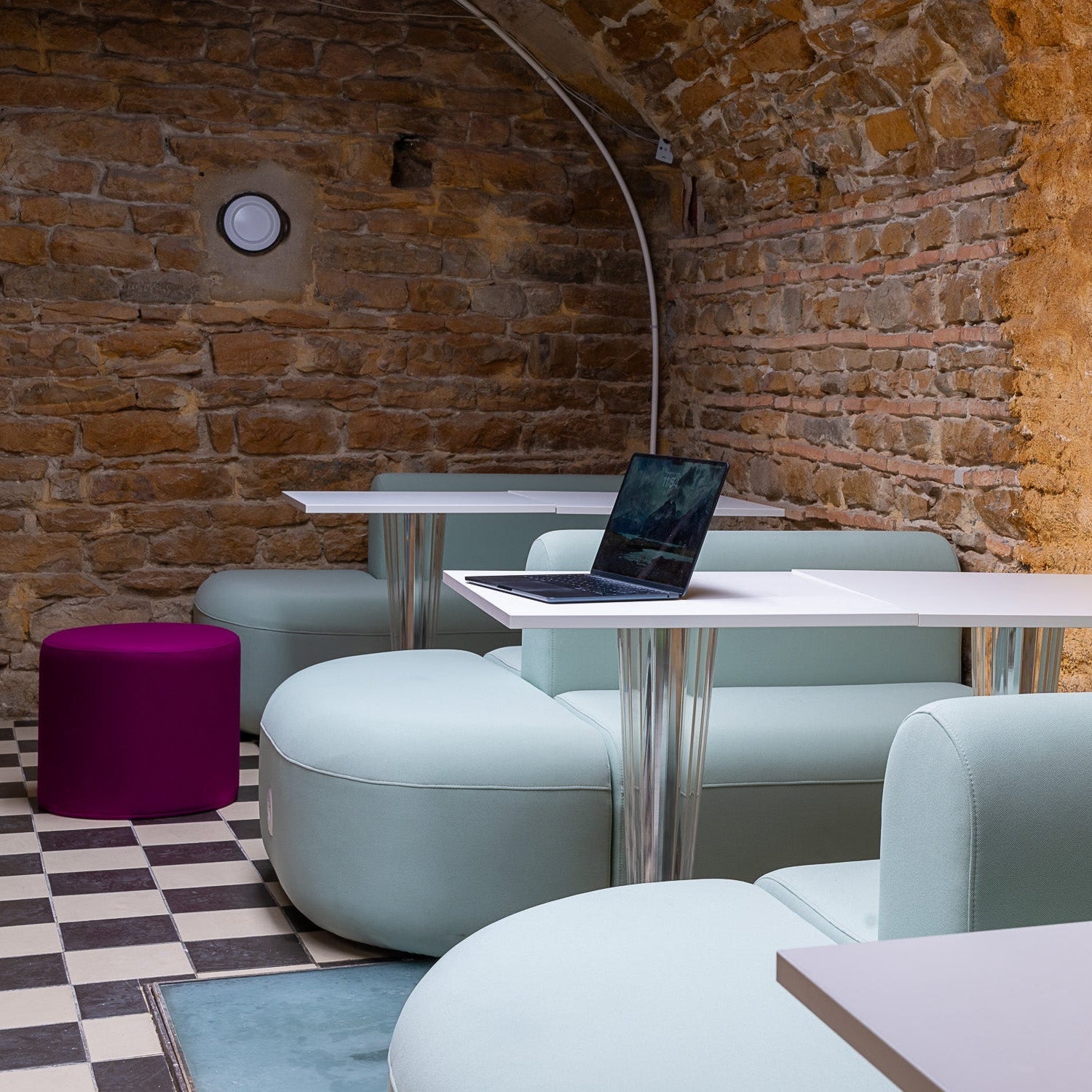 Modern interior with light blue chairs, a purple stool, and a laptop on a table against a brick wall.