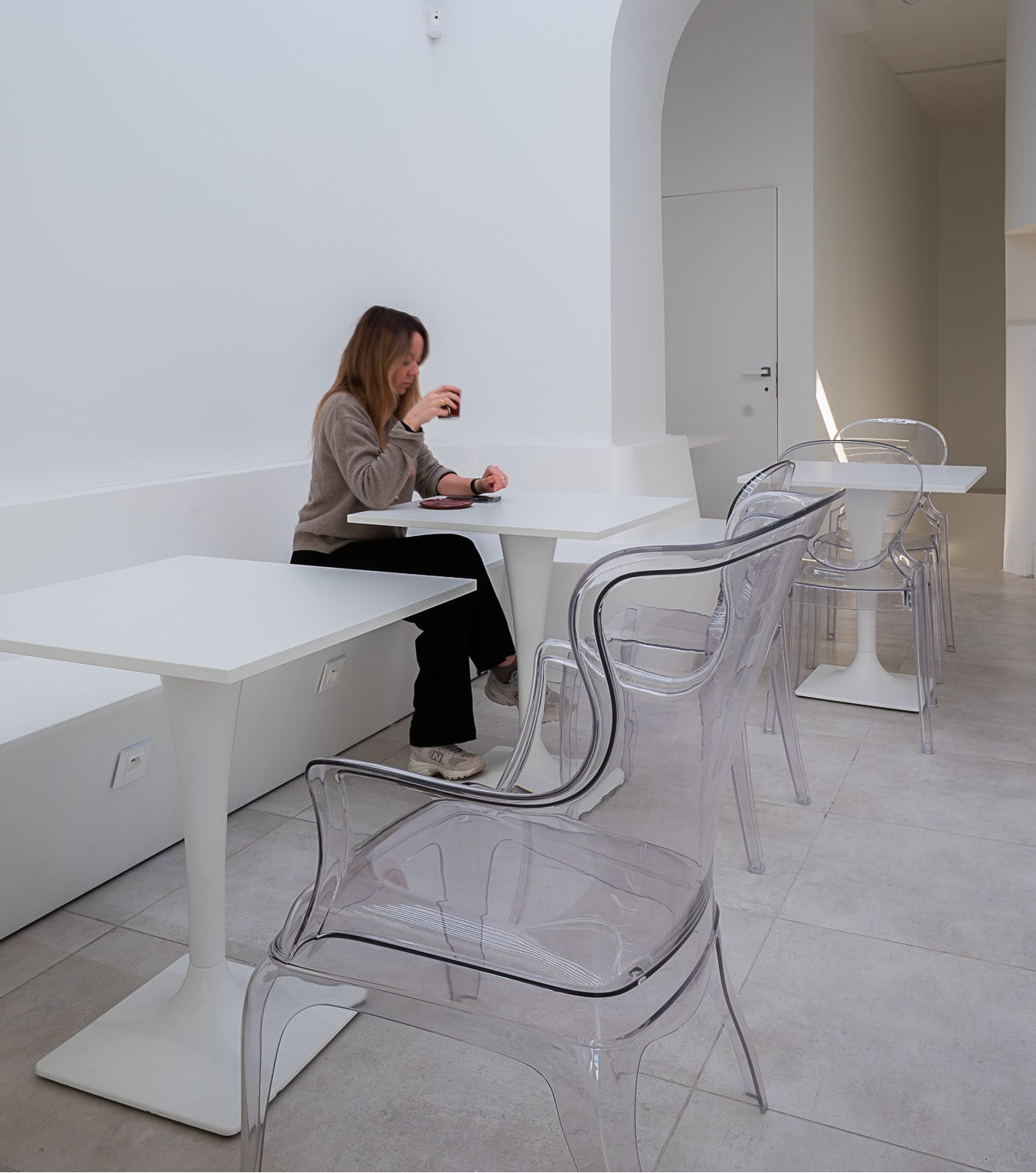 Woman sitting at a white table in a minimalistic room with clear chairs.