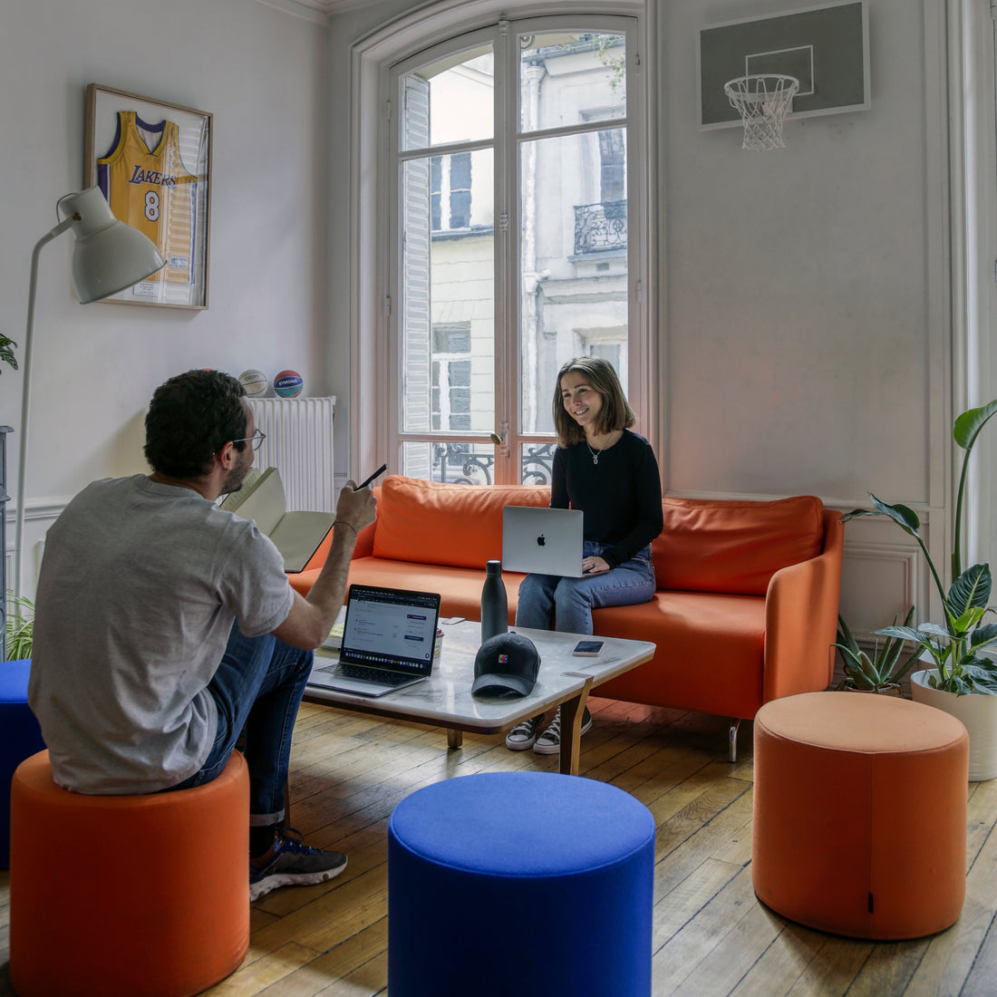 Two people sitting on orange and blue stools in a modern relaxed office meeting room with orange sofa, blue and orange poufs and basketball hoop.