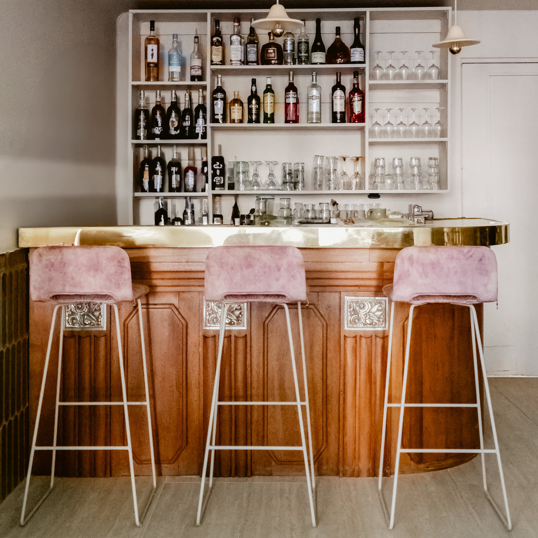 Modern bar with wooden counter, pink stools, and shelves stocked with bottles and glasses.
