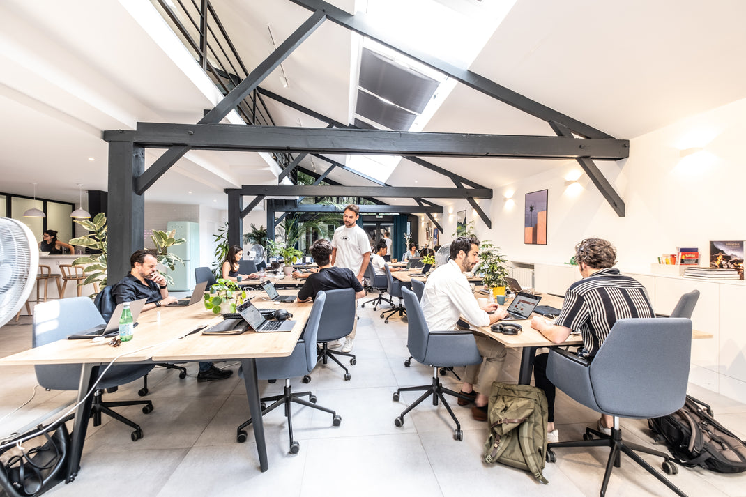 Modern office space with people working at desks, large windows, and exposed beams.