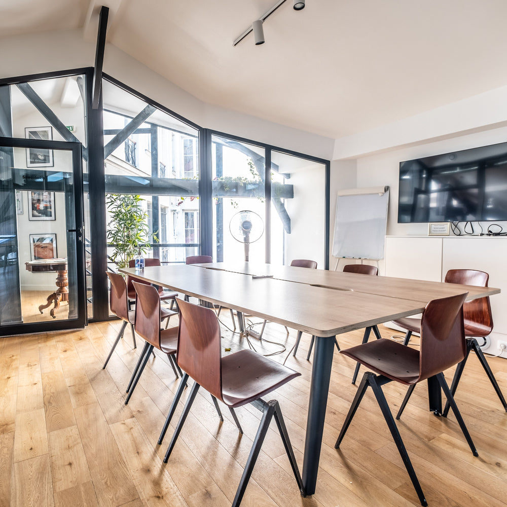 Modern office space with a long wooden table and chairs, large windows, and a TV on the wall.