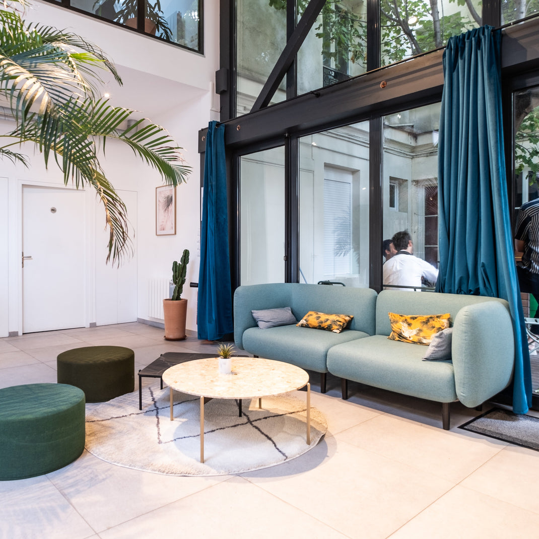Modern lounge area in office open plan with a light blue sofa, green ottomans, and a coffee table in a room with large windows.